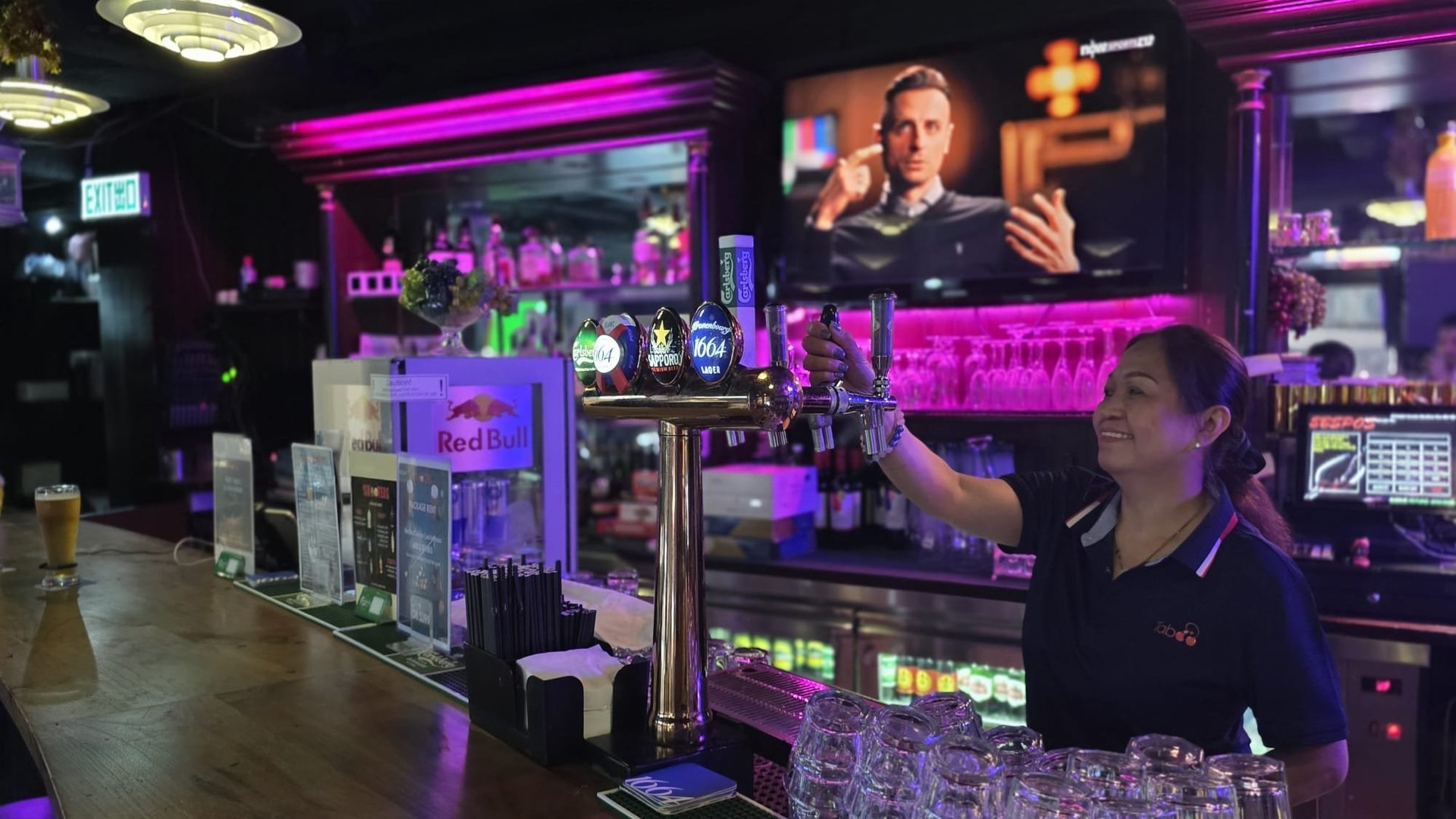 Bartender at a nightclub bar counter lit with purple and pink neon lighting, with bottles displayed and screens on the walls