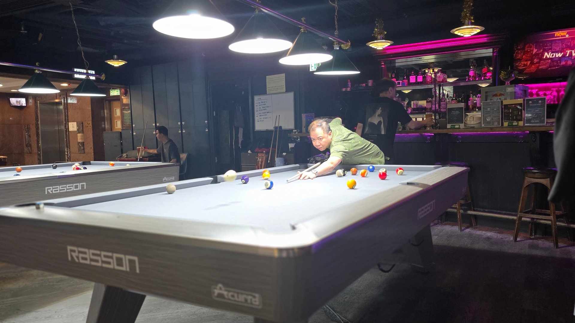 Person playing pool at an Airson billiards table in a dimly lit arcade venue with neon signage and overhead lighting